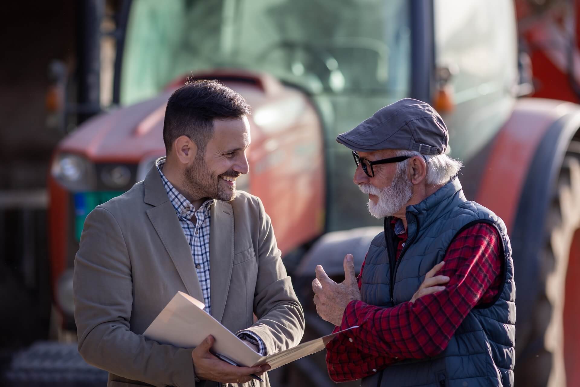 Business agent and senior farmer talking about loan contract with bank in front of tractor on ranch